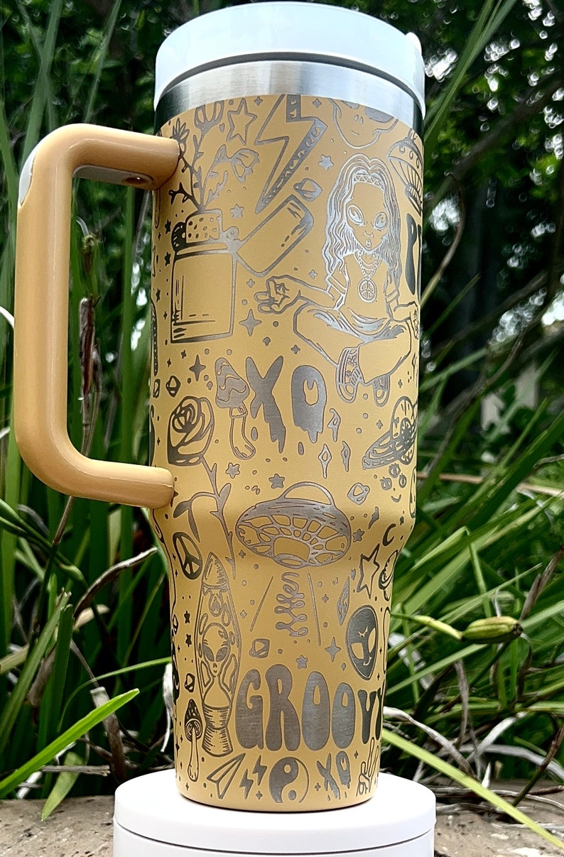 May include: A beige travel mug with a handle, featuring etched illustrations of aliens, mushrooms, a peace sign, and the word "GROOVY." The mug has a white lid and a stainless steel rim.