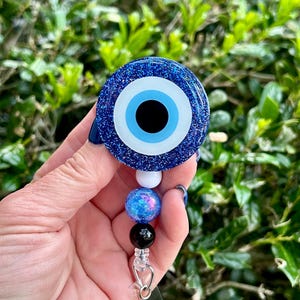 May include: A circular badge reel featuring a dark blue glittery evil eye design with a central black pupil and light blue iris. Below the circular piece, a vertical strand of three beads in white, blue glitter, and black connects to a metal clip.