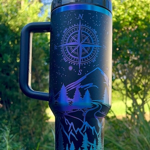 May include: A black stainless steel travel mug with a handle and a straw. The mug has a compass design with a mountain scene and trees. The mug is on a white coaster.