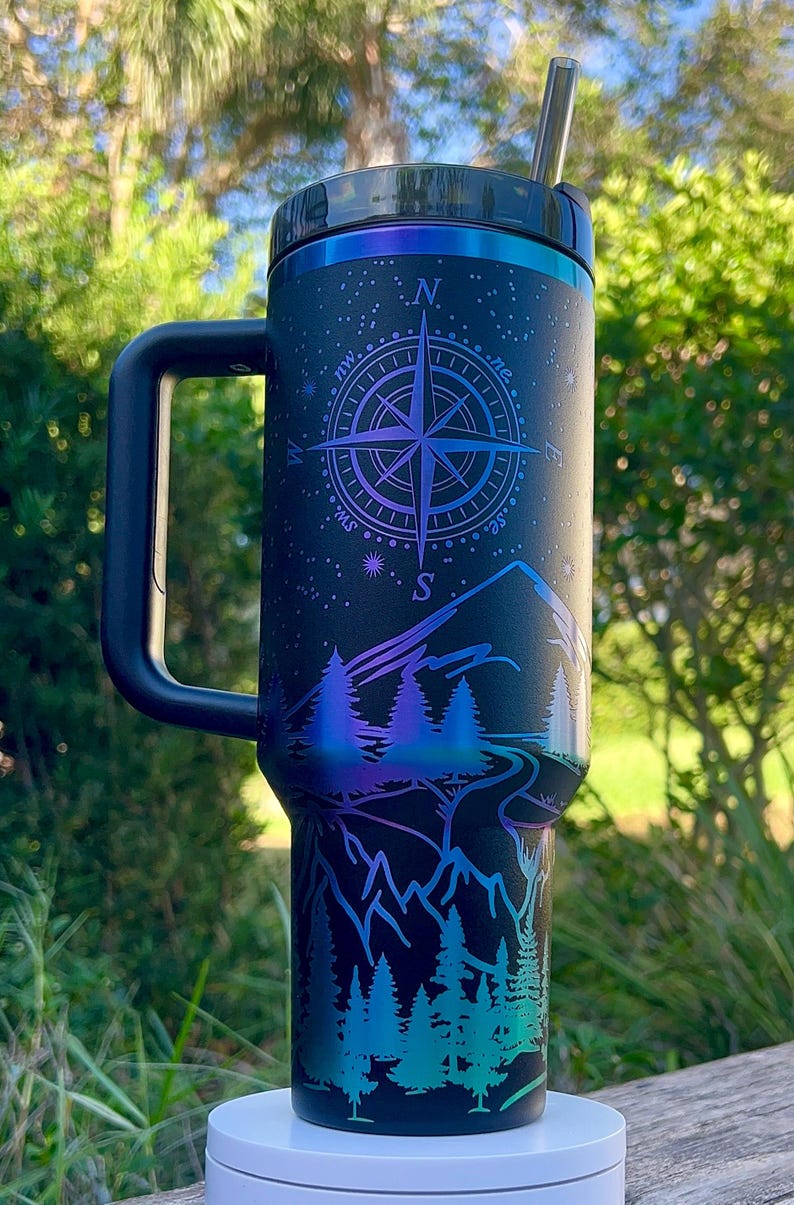 May include: A black stainless steel travel mug with a handle and a straw. The mug has a compass design with a mountain scene and trees. The mug is on a white coaster.
