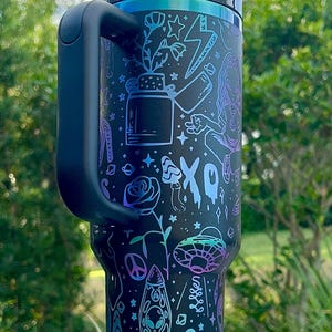 May include: Black tumbler with a handle, featuring a colorful design of plants, stars, an alien, and the word "GROOVE". The tumbler has a black lid and a straw. The design includes shades of blue, purple, and green.