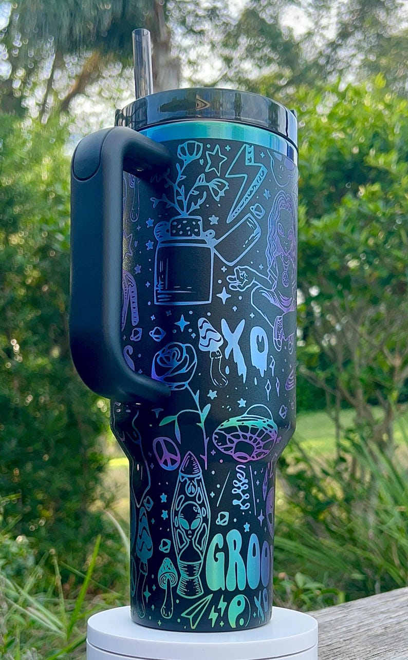 May include: Black tumbler with a handle, featuring a colorful design of plants, stars, an alien, and the word "GROOVE". The tumbler has a black lid and a straw. The design includes shades of blue, purple, and green.