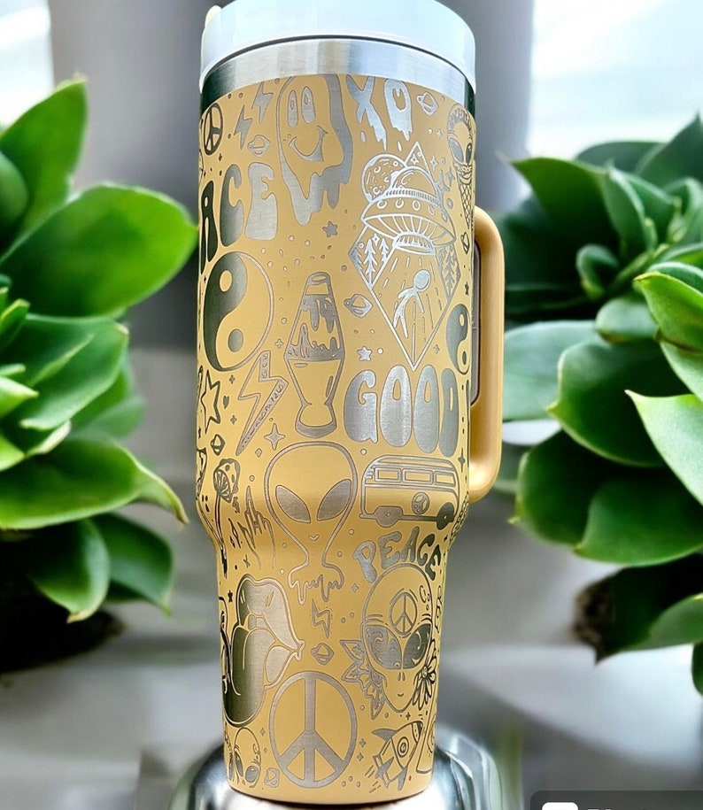 May include: A yellow insulated tumbler with a white lid and handle. The tumbler is covered in etched designs including aliens, peace signs, a lava lamp, and the words "Peace" and "Good".