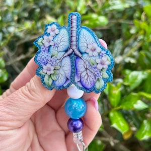 May include: A blue glitter badge reel featuring a stylized illustration of lungs adorned with purple and white flowers. The badge reel has a beaded design with white, blue, and purple beads.