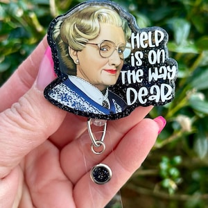 Puede incluir: Un portagafete negro con purpurina con una imagen de una mujer con el texto "Help is on the way dear".