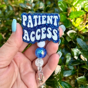 May include: A blue glitter badge reel with the words "PATIENT ACCESS" in white lettering. The badge reel has a beaded design with a white, blue, and clear bead. The background is blurred green foliage.