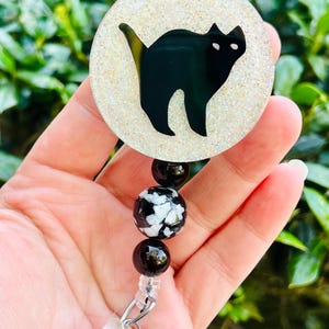 Puede incluir: Un portatarjetas circular con la silueta de un gato negro sobre un fondo blanco brillante. El portatarjetas est&aacute; unido a un segmento de cuentas con dos cuentas negras s&oacute;lidas y una cuenta central con un patr&oacute;n de m&aacute;rmol en blanco y negro.