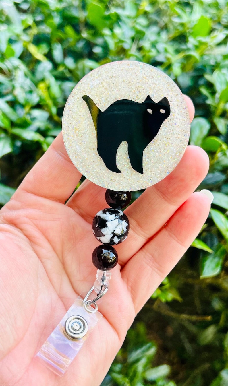 Puede incluir: Un portatarjetas circular con la silueta de un gato negro sobre un fondo blanco brillante. El portatarjetas est&aacute; unido a un segmento de cuentas con dos cuentas negras s&oacute;lidas y una cuenta central con un patr&oacute;n de m&aacute;rmol en blanco y negro.