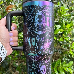 May include: Black travel mug with a handle, featuring a colorful alien-themed design. The design includes illustrations of aliens, UFOs, planets, and the word "Groovy". The mug has a black lid and handle, and a gradient of blue, purple, and pink colors.
