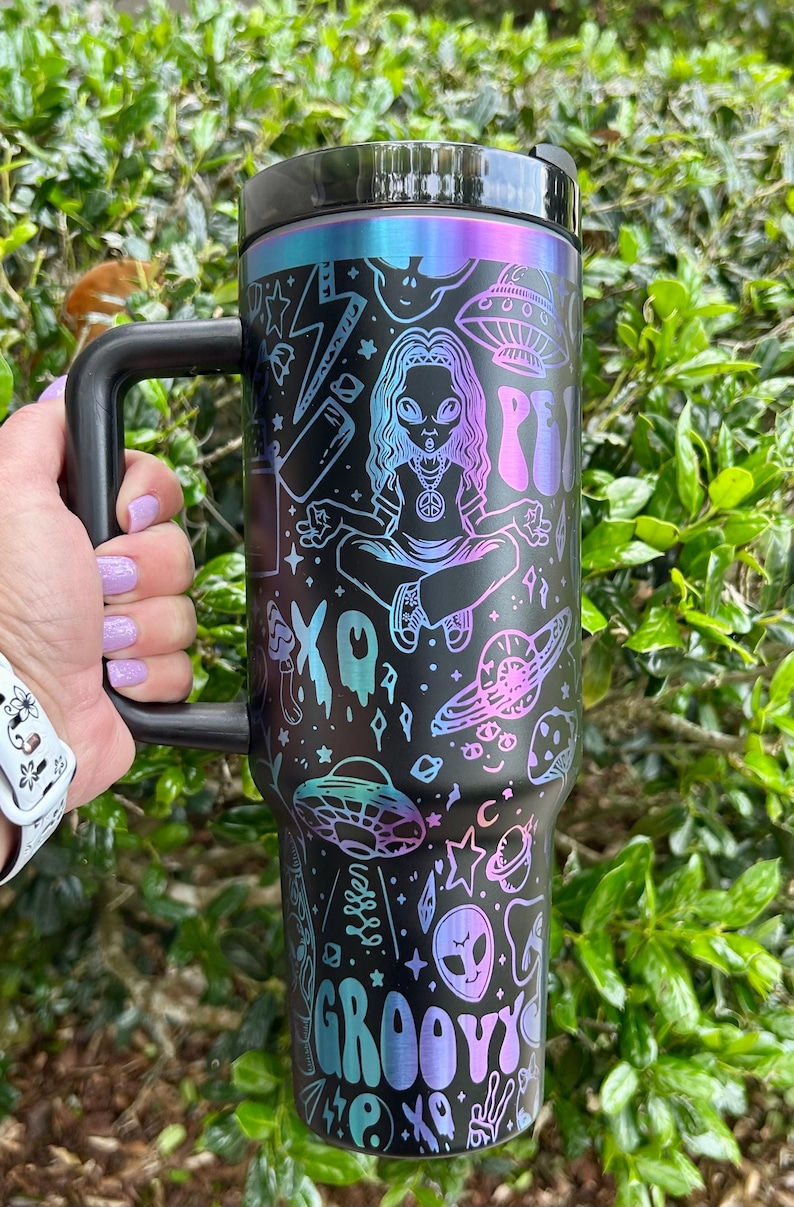 May include: Black travel mug with a handle, featuring a colorful alien-themed design. The design includes illustrations of aliens, UFOs, planets, and the word "Groovy". The mug has a black lid and handle, and a gradient of blue, purple, and pink colors.