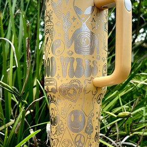 May include: A yellow travel mug with a white lid and handle, featuring etched designs of aliens, peace signs, and the words "GOOD VIBES." The mug is cylindrical and stands on a white base, perfect for carrying beverages on the go.
