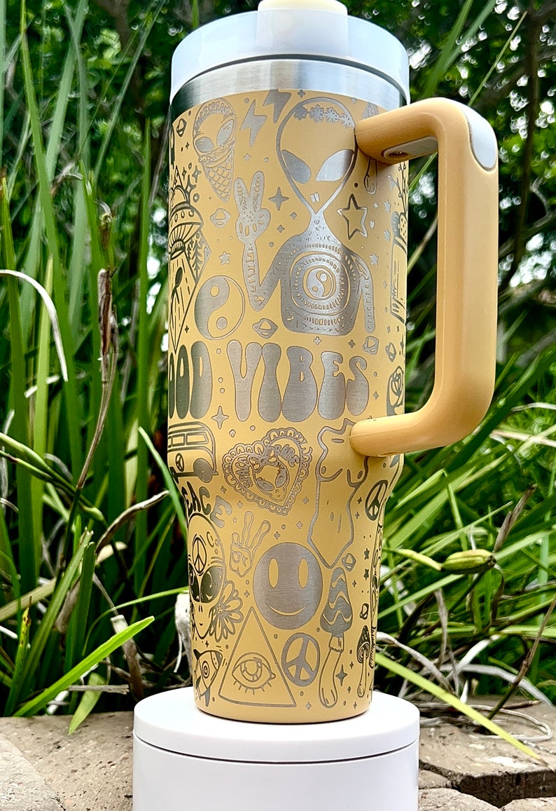 May include: A yellow travel mug with a white lid and handle, featuring etched designs of aliens, peace signs, and the words "GOOD VIBES." The mug is cylindrical and stands on a white base, perfect for carrying beverages on the go.