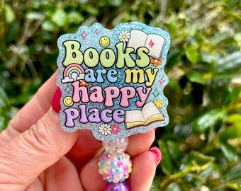 Puede incluir: Un portatarjetas con un adorno acrílico azul brillante en forma de nube con el texto Books are my happy place en letras de burbuja coloridas. El diseño incluye ilustraciones de libros abiertos, un arcoíris, flores y caras sonrientes.