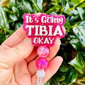 May include: Pink glitter badge holder with a pink and white swirl bead and a pink glitter bead. The badge holder has a clear plastic clip and the text "It's Going Tibia Okay" on the front.