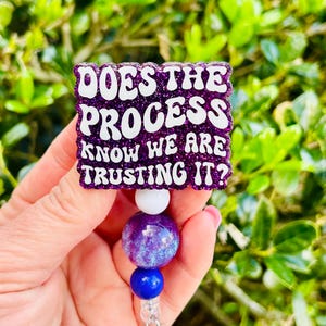 May include: A purple glitter badge reel with the text "DOES THE PROCESS KNOW WE ARE TRUSTING IT?" in white. The reel has a white, purple, and blue beaded design.