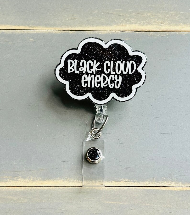 以下が含まれることがあります： 黒いグリッター仕上げの雲型バッジリールで、白い文字で「BLACK CLOUD ENERGY」とスタイリッシュなフォントで書かれています。バッジリールには、金属製のスナップボタンが付いた透明なプラスチックストラップと、身分証を保持するための伸縮式コード機構が含まれています。