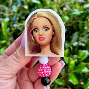 Puede incluir: Un portatarjetas con la cara de una muñeca con expresión de sorpresa. El portatarjetas tiene un diseño de cuentas blancas, rosas y negras. La muñeca tiene pelo rubio, ojos grandes y pintalabios rosa.