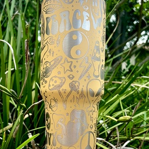 May include: A beige stainless steel tumbler with a white lid, featuring etched designs of peace signs, mushrooms, aliens, and the word "PEACE". The tumbler has a silver rim and a white base, perfect for cold beverages.