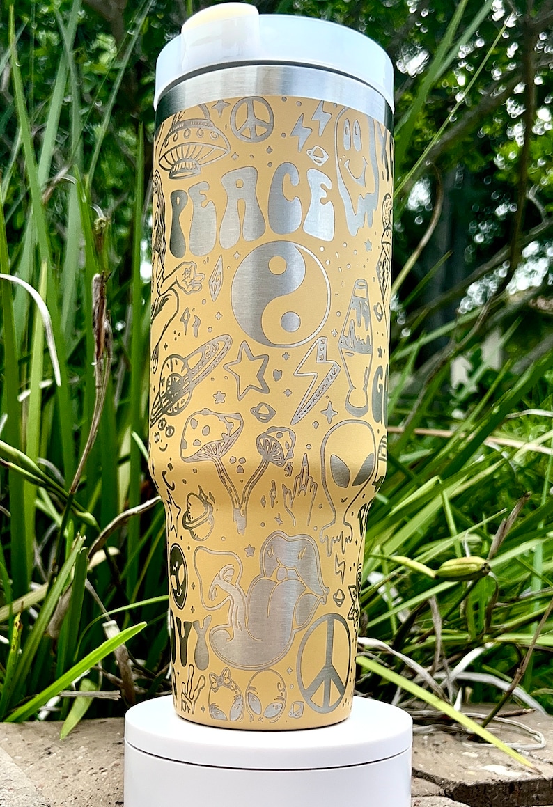 May include: A beige stainless steel tumbler with a white lid, featuring etched designs of peace signs, mushrooms, aliens, and the word "PEACE". The tumbler has a silver rim and a white base, perfect for cold beverages.