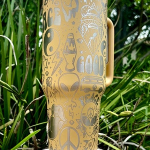 May include: A beige travel mug with a white lid and handle, adorned with etched silver designs. The design includes a smiley face, alien, peace signs, and the word "GOOD". The mug is on a white stand.