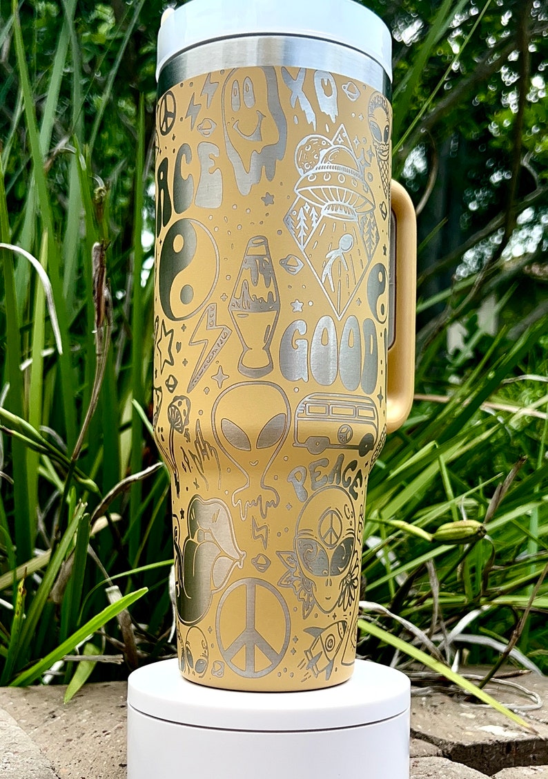 May include: A beige travel mug with a white lid and handle, adorned with etched silver designs. The design includes a smiley face, alien, peace signs, and the word "GOOD". The mug is on a white stand.
