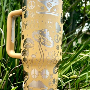 May include: A yellow stainless steel travel mug with a silver lid and handle. The mug features a peace sign, mushrooms, flowers, and a smiley face design.
