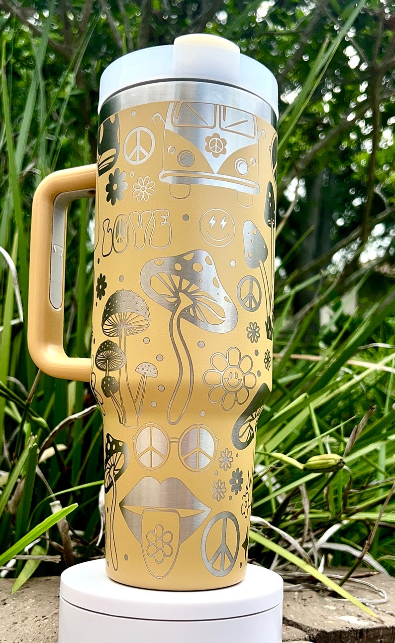 May include: A yellow stainless steel travel mug with a silver lid and handle. The mug features a peace sign, mushrooms, flowers, and a smiley face design.