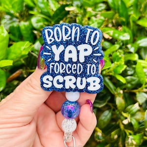 May include: A blue glitter badge reel with the text "BORN TO YAP FORCED TO SCRUB" in white. The badge reel has a beaded design with a white, blue, and clear bead. The background is blurred green foliage.