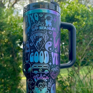 May include: Black Stanley tumbler with a handle, featuring a colorful, psychedelic design. The design includes the text "GOOD VIBES" and images of aliens, a peace sign, a VW bus, and a yin-yang symbol. The tumbler has a black lid and a silver straw.