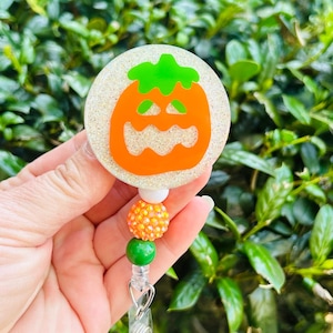 Puede incluir: Un portatarjetas circular con fondo blanco brillante y un gr&aacute;fico de calabaza naranja. El carrete est&aacute; unido a una hilera vertical de cuentas que incluye una blanca, una naranja con textura y una cuenta verde lisa.