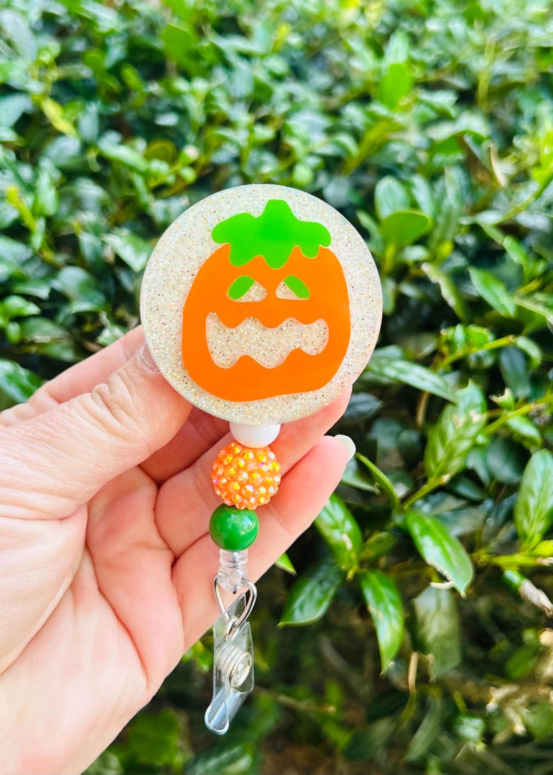 Puede incluir: Un portatarjetas circular con fondo blanco brillante y un gr&aacute;fico de calabaza naranja. El carrete est&aacute; unido a una hilera vertical de cuentas que incluye una blanca, una naranja con textura y una cuenta verde lisa.