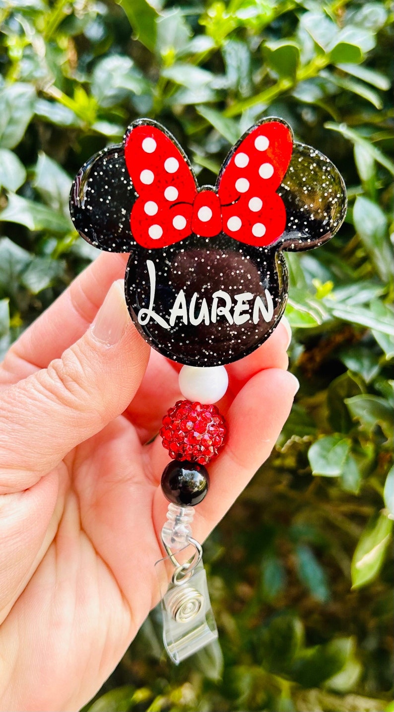 May include: A black and red glitter Minnie Mouse themed badge holder with the name "LAUREN" on the front. The holder has a red and black beaded chain with a white bead and a red rhinestone bead.