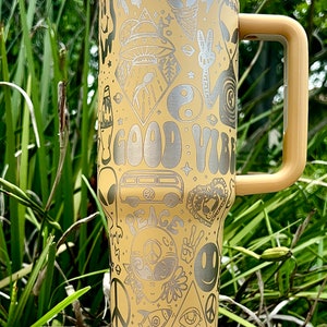 May include: A beige travel mug with a white lid and a tan handle. The mug is covered in etched designs, including aliens, a peace sign, a smiley face, and the words "GOOD VIBES" and "PEACE".