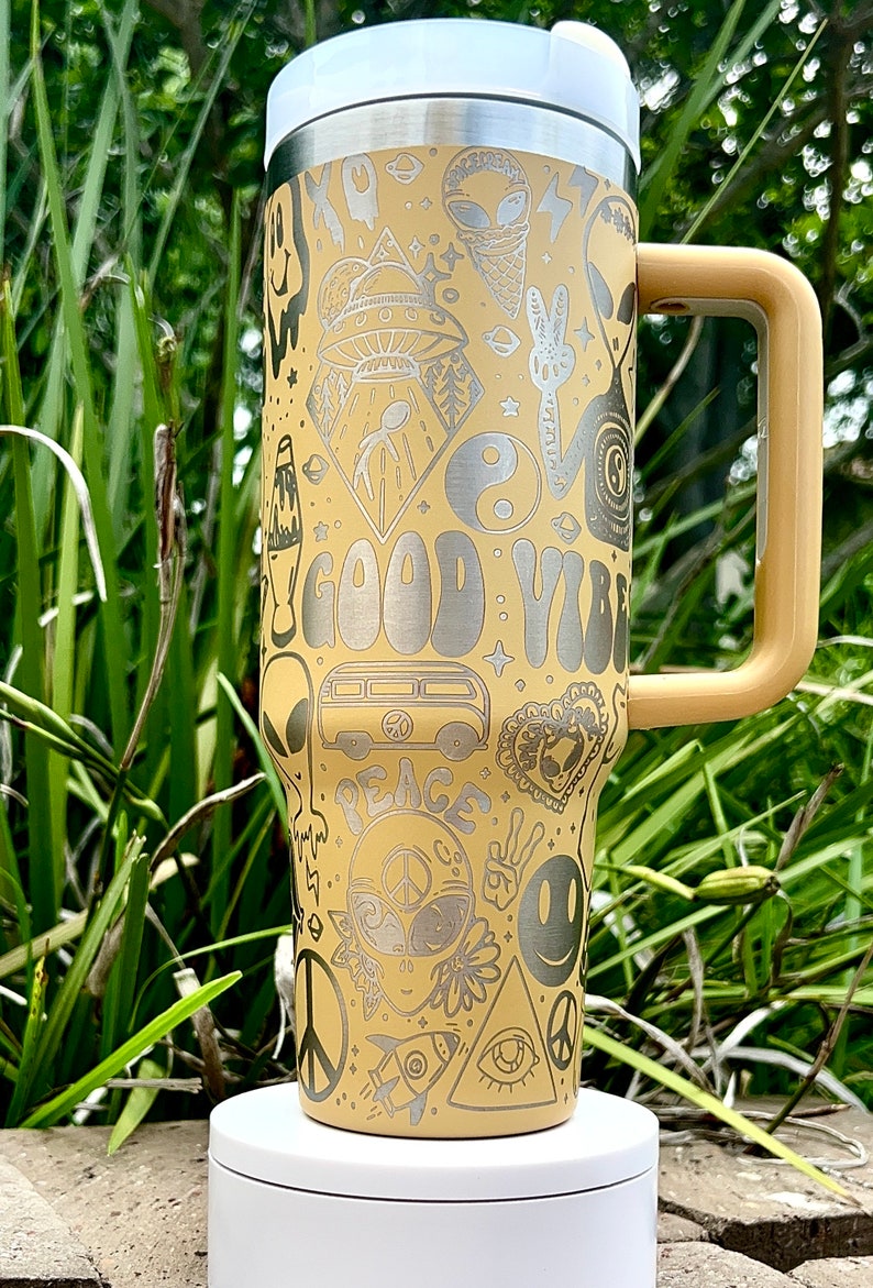May include: A beige travel mug with a white lid and a tan handle. The mug is covered in etched designs, including aliens, a peace sign, a smiley face, and the words "GOOD VIBES" and "PEACE".