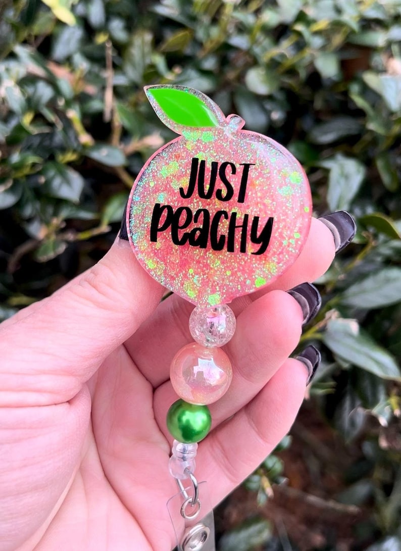 May include: A pink peach-shaped badge holder with glitter and the text "Just Peachy". The holder has a green leaf and is attached to a clear retractable badge reel with three beads: a clear bead, a pink iridescent bead, and a green bead.