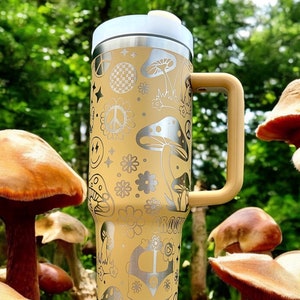 May include: A yellow stainless steel travel mug with a black handle. The mug is decorated with a psychedelic pattern of mushrooms, peace signs, and flowers. The mug is sitting on a wooden surface with mushrooms in the background.