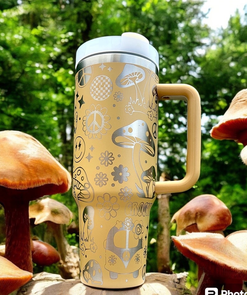 May include: A yellow stainless steel travel mug with a black handle. The mug is decorated with a psychedelic pattern of mushrooms, peace signs, and flowers. The mug is sitting on a wooden surface with mushrooms in the background.
