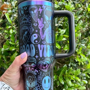 May include: Black travel mug with a black handle and lid, featuring a colorful design with the words "GOOD VIBES" and "PEACE". The design includes aliens, ice cream cones, peace signs, and other psychedelic graphics.