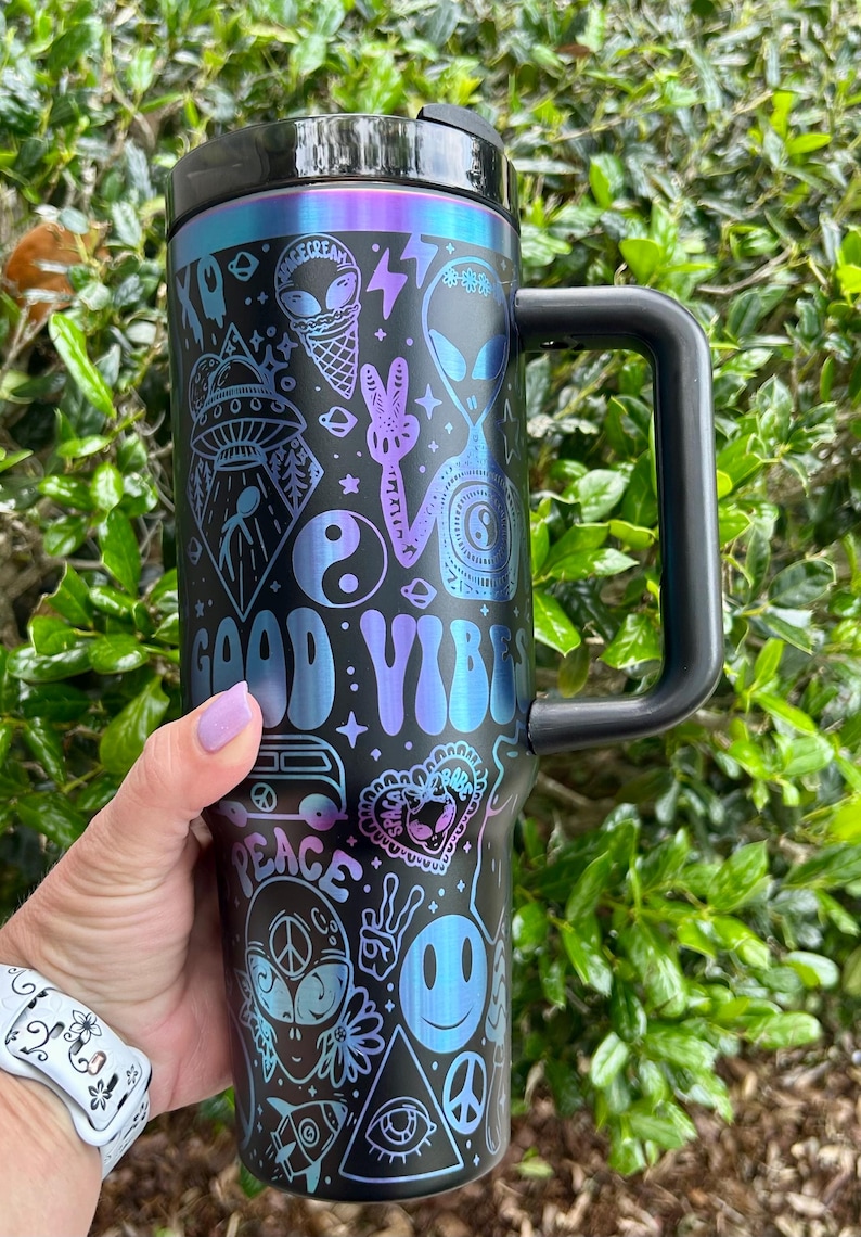 May include: Black travel mug with a black handle and lid, featuring a colorful design with the words "GOOD VIBES" and "PEACE". The design includes aliens, ice cream cones, peace signs, and other psychedelic graphics.