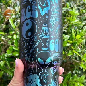 May include: Black and blue insulated tumbler with a variety of psychedelic-themed graphics. The design includes a peace sign, an alien, a lava lamp, and the word "GOOD" in a groovy font. The tumbler has a black lid.
