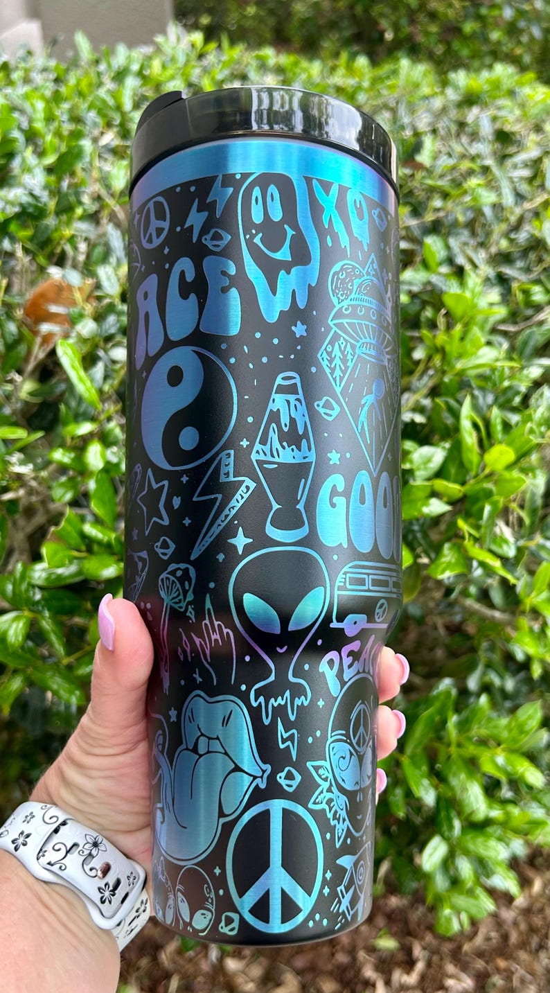 May include: Black and blue insulated tumbler with a variety of psychedelic-themed graphics. The design includes a peace sign, an alien, a lava lamp, and the word "GOOD" in a groovy font. The tumbler has a black lid.