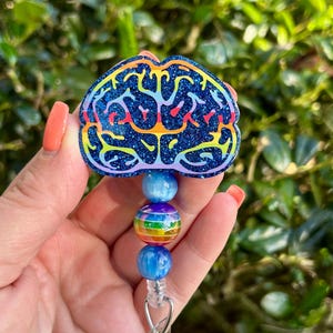 May include: A blue glitter badge reel with a brain design. The brain is outlined in rainbow colors. The badge reel has blue and rainbow-colored beads. The badge reel is attached to a silver clip.