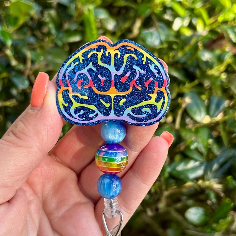 May include: A blue glitter badge reel with a brain design. The brain is outlined in rainbow colors. The badge reel has blue and rainbow-colored beads. The badge reel is attached to a silver clip.