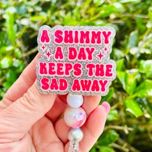May include: A glittery badge reel with the text "A Shimmy A Day Keeps The Sad Away" in pink lettering. The badge has a silver glitter border and is attached to a beaded lanyard with white, pink, and silver beads.