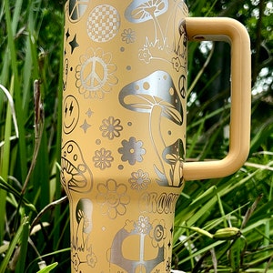 May include: A yellow stainless steel travel mug with a white lid and a brown handle. The mug is decorated with a psychedelic pattern of peace signs, mushrooms, flowers, and a van. The text "Groovy" is also visible on the mug.