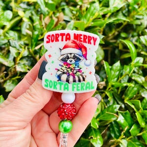 May include: A badge reel with a raccoon wearing a Santa hat and Christmas lights. The text on the badge reads "Sorta Merry" and "Sorta Feral" in red and green. The reel has a white bead, a red beaded section, and a green bead.