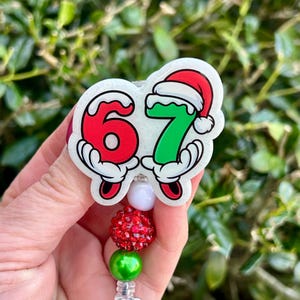 May include: A festive badge reel featuring the numbers 67 in red and green, each held by cartoon hands. The number 7 is wearing a Santa hat. The reel has red and green beaded accents.