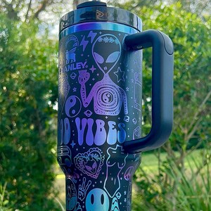 May include: Black and teal insulated tumbler with a handle and a straw. The tumbler features a design with aliens, peace signs, mushrooms, and the words "Good Vibes" in a gradient of teal and purple.