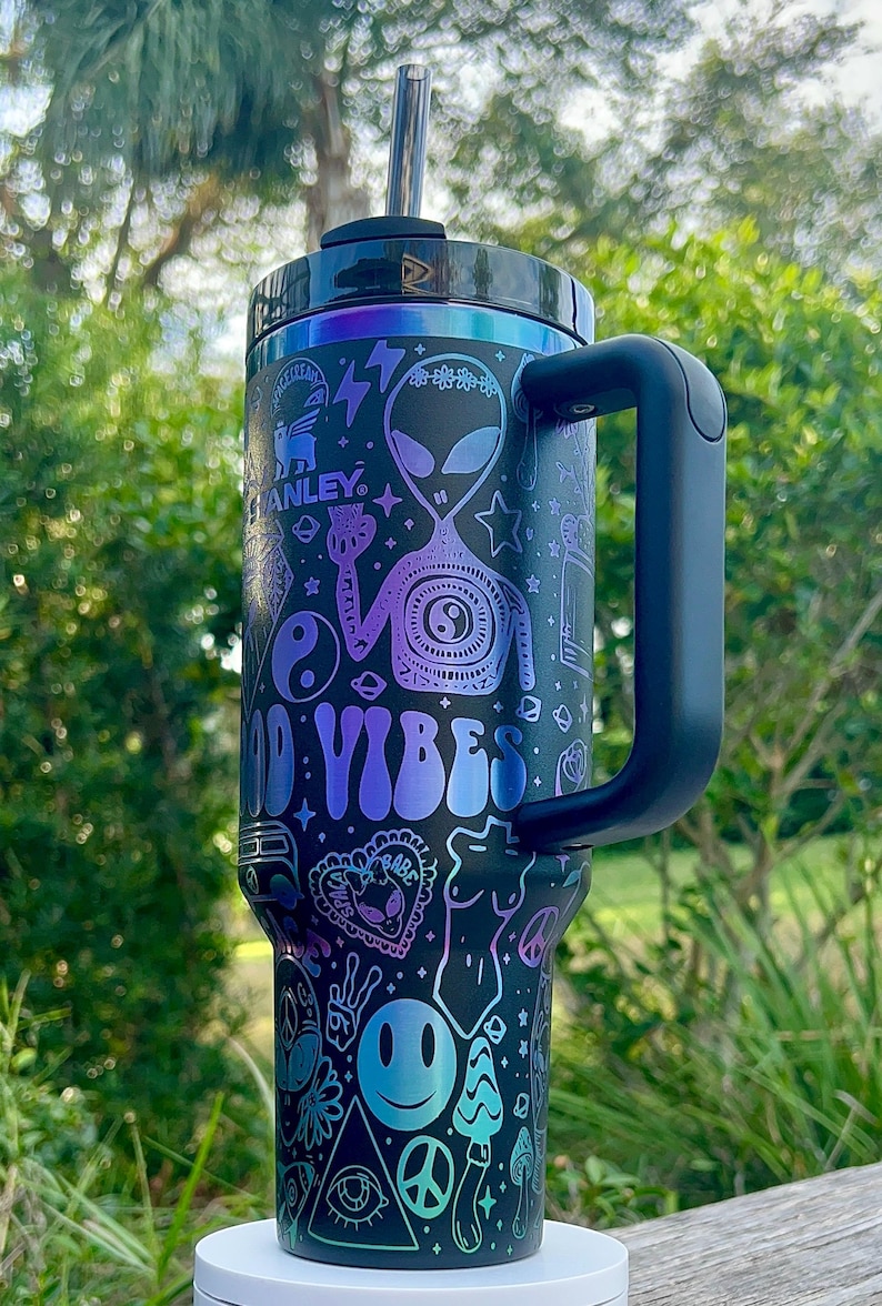 May include: Black and teal insulated tumbler with a handle and a straw. The tumbler features a design with aliens, peace signs, mushrooms, and the words "Good Vibes" in a gradient of teal and purple.
