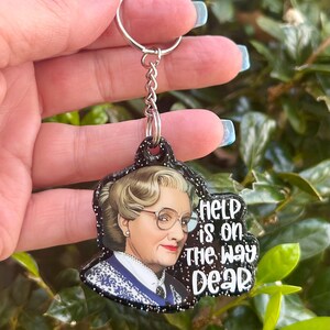 Puede incluir: Un llavero de purpurina negra con una imagen de una mujer con cabello rubio y gafas. El texto en el llavero dice "Help is on the way dear."
